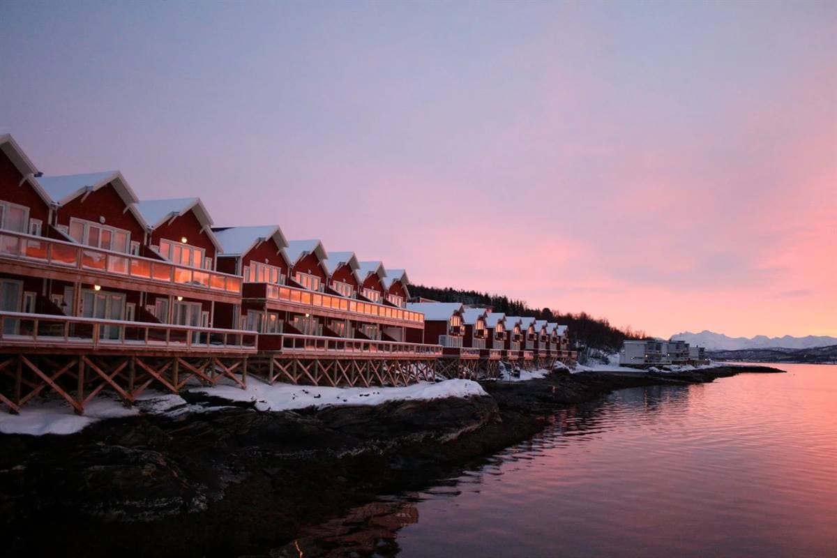 Long red two-storey building on stilts over the calm fjord at winter dusk