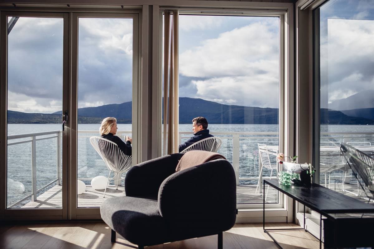 Two guests on a balcony overlooking the fjord, seen from inside a cabin