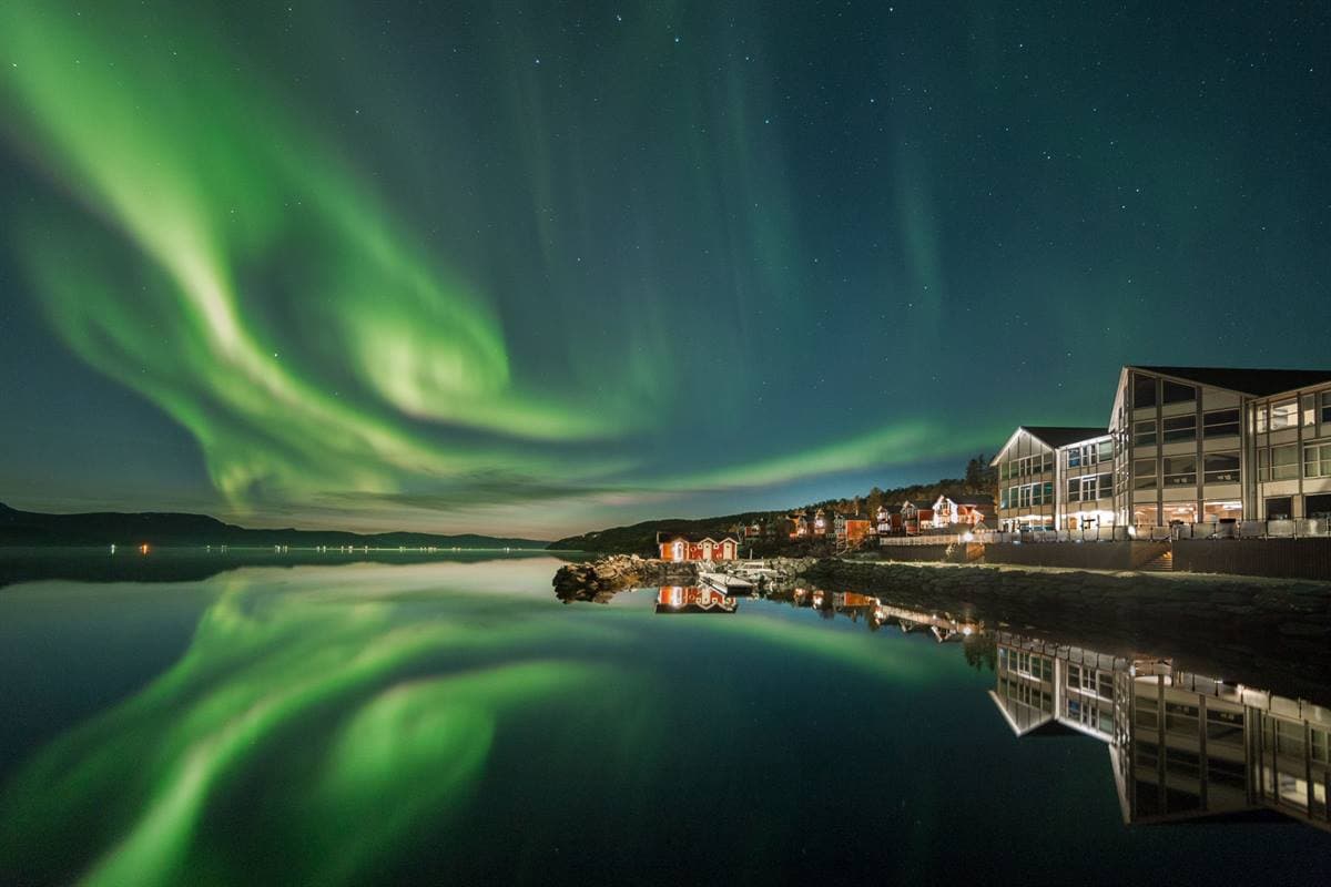 Northern lights over the Malangen fjord, reflected in the water beside the lit-up resort